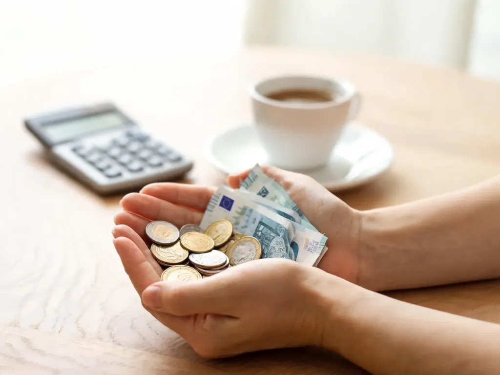 Dłonie trzymające monety i banknoty euro na stole, w tle kalkulator i kawa, symbolizujące analizę najniższej krajowej w Hiszpanii.